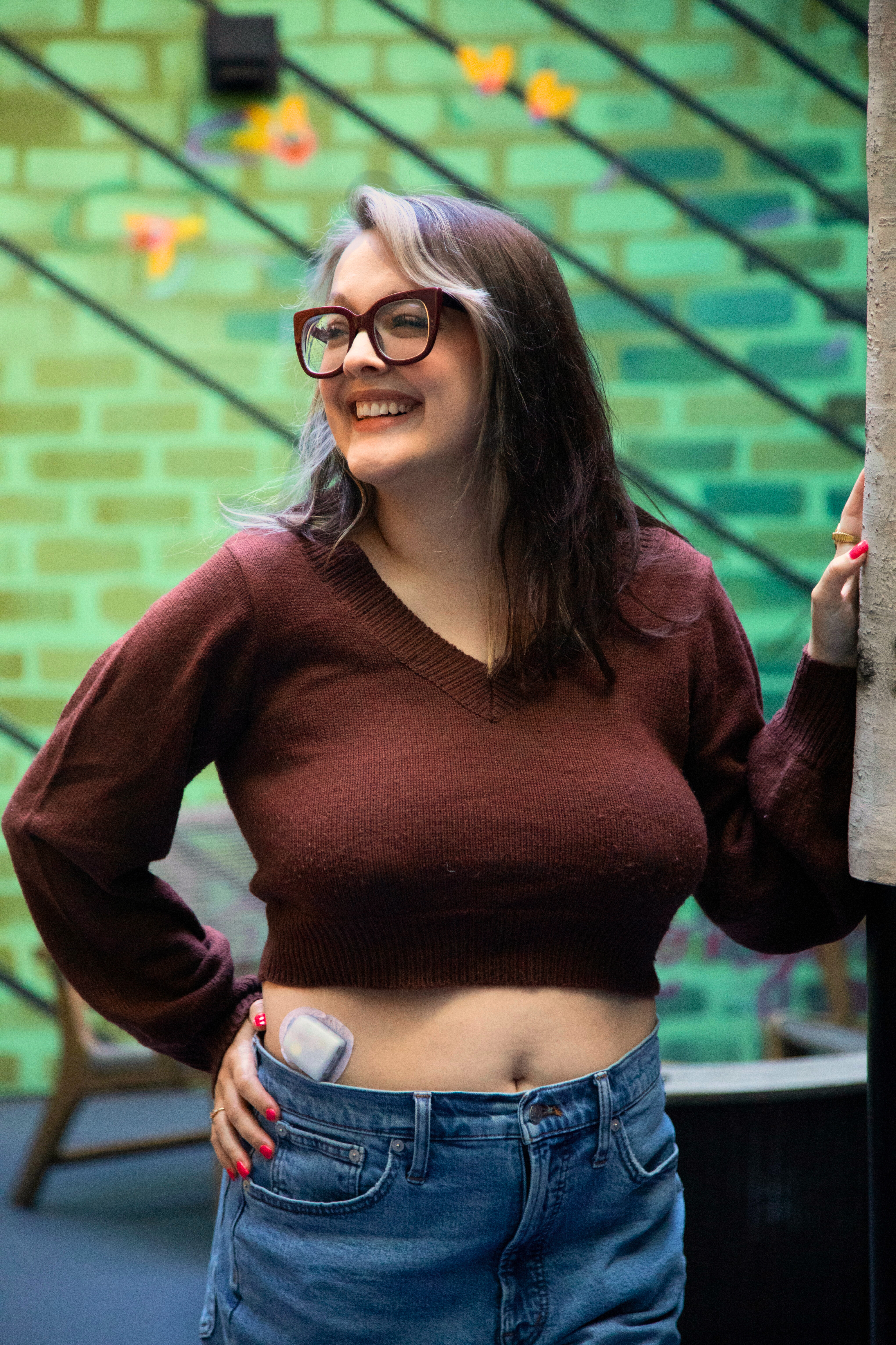 A smiling woman wearing glasses stands with her hand on her hip, showcasing a cropped sweater and high-waisted jeans, against a colorful green brick wall with decorative butterflies.